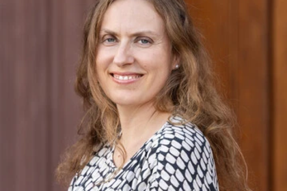 Die Person hat lange, lockige Haare, trägt ein schwarz-weiß-gemustertes Oberteil und lächelt.