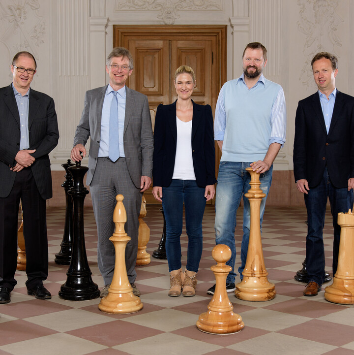 Fünf Personen in schicker Kleidung stehen auf einem Schachfeld.