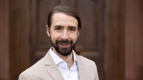 Johannes Stäbler. He is wearing a beige suit and is standing in front of brown door.