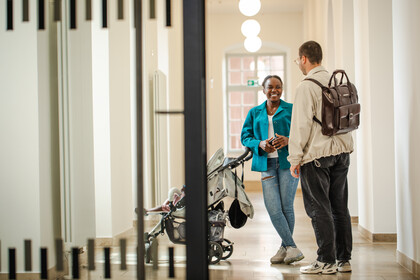 Zwei Forschende stehen im Flur des Ostflügels und unterhalten sich lächelnd. Neben ihnen steht ein Kinderwagen.