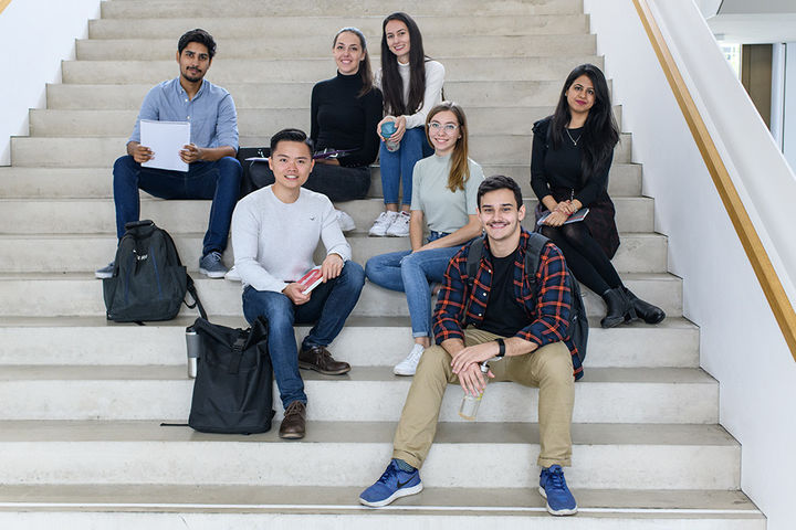 Eine Gruppe von sieben Studierenden sitzt auf einer hellen Treppe und lächelt in die Kamera. Sie haben Rucksäcke und Notizblöcke bei sich.