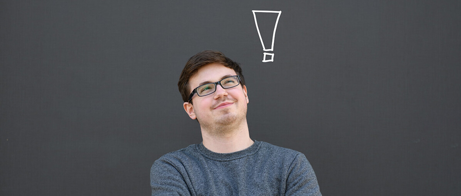 A young man is standing in front of a grey background. He is looking up and above his head is a exclamation mark.