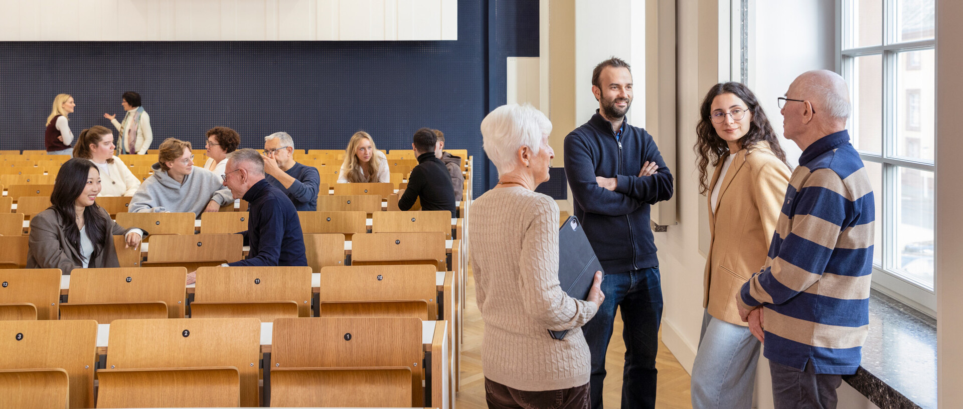 Im Vordergrund unterhalten sich vier Menschen unterschiedlichen Alters. Im Hintergrund unterhalten sich weitere Menschen sitzend und stehend.