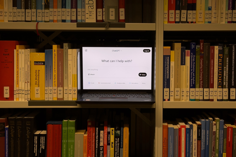 In einem Bücherregal ist ein aufgeklappter Laptop auf dem ein ChatGPT-Fenster geöffnet ist.