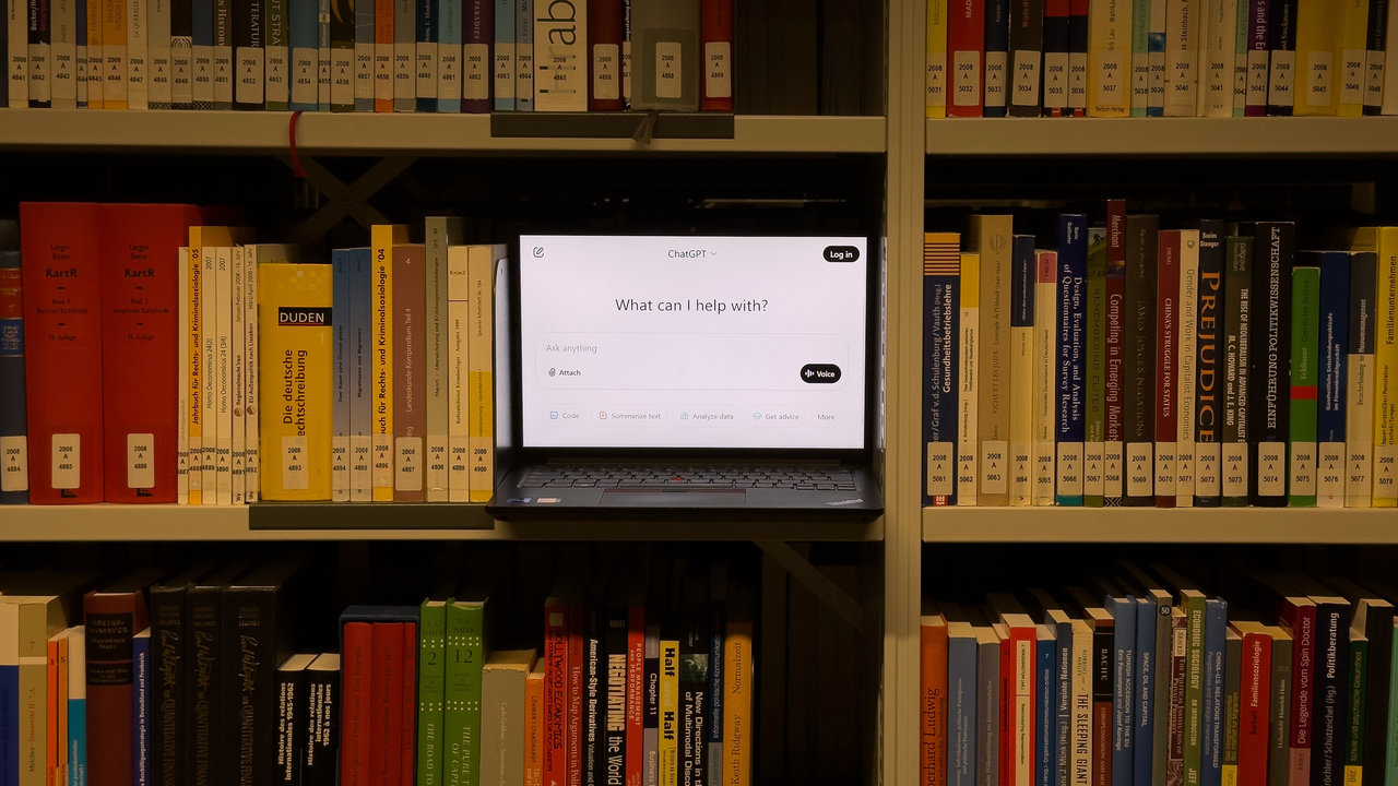 In einem Bücherregal ist ein aufgeklappter Laptop auf dem ein ChatGPT-Fenster geöffnet ist.