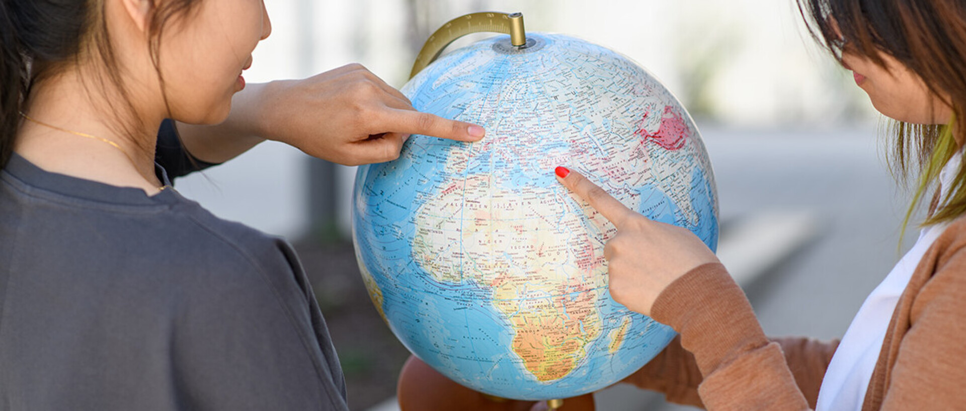 Two women are holding a globe. The one on the left is pointing at Mannheim and the one on the right is pointing at a country in the Near East.