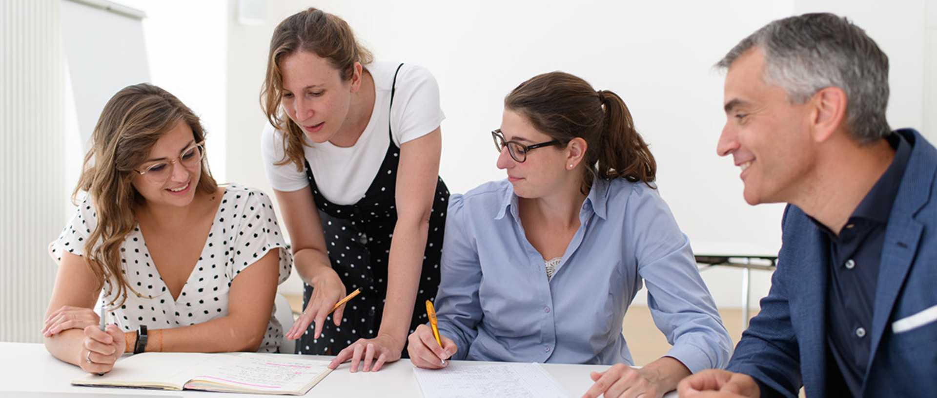 Zwei Forscherinnen und ein Forscher sitzen am Tisch, während eine Forscherin stehend Aufgeschriebens in einem Notizbuch erklärt
