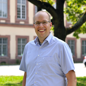 Alexander Pfister hat kurze graue Haare und eine Brille. Er trägt ein helles, kurzärmeliges Hemd und lächelt freundlich.