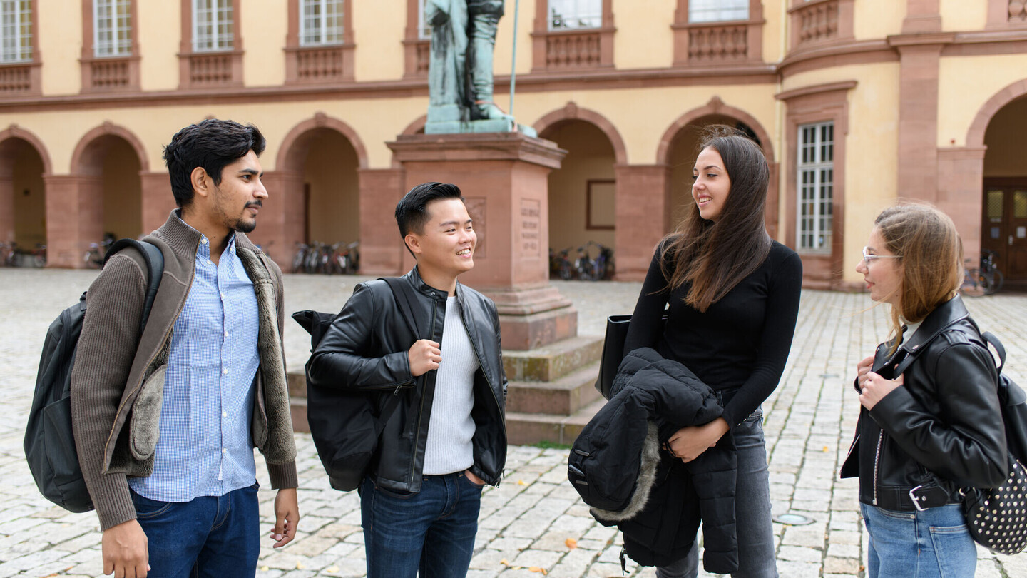 Eine Gruppe von 4 Studierenden steht vor einer Statue, im Hintergrund das Barockschloss.