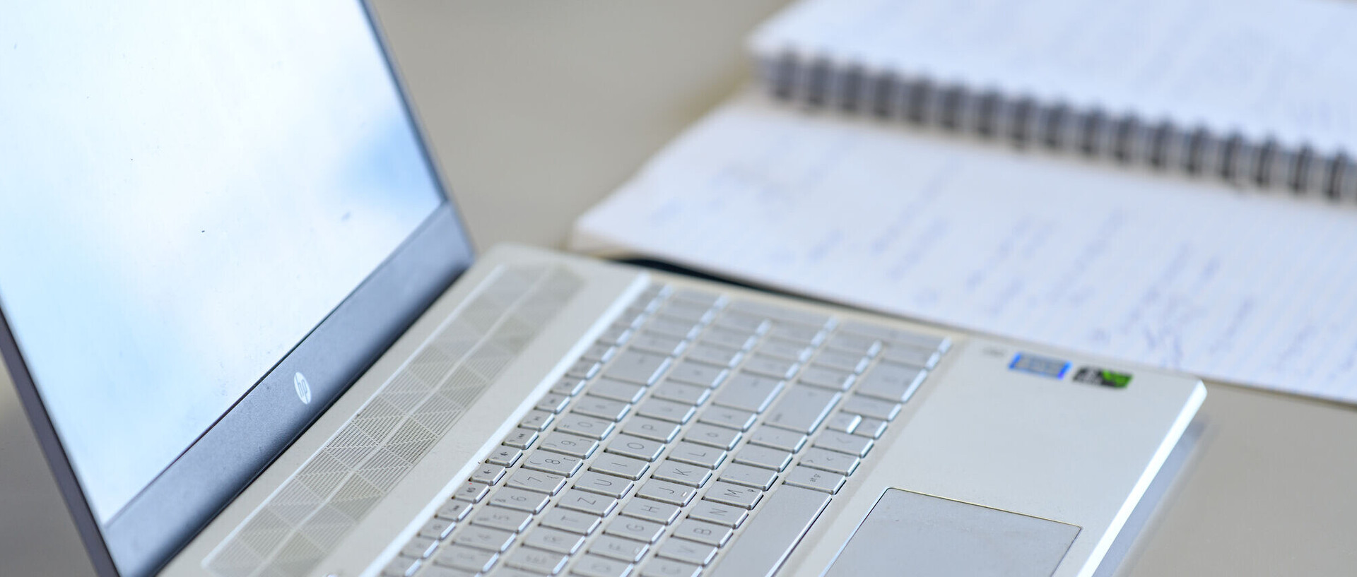 Ein aufgeklappter Laptop steht auf dem Tisch, daneben liegt ein beschriebenes Notizbuch.