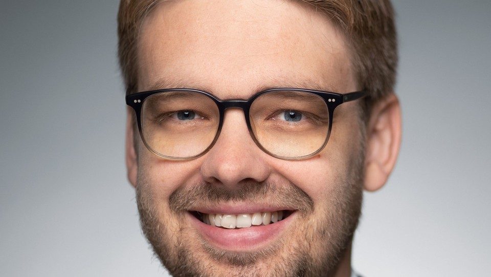 Eine Person trägt ein weißes Hemd sowie einen schwarzen Blazer und steht vor einer grauen Wand. Die Person heißt Marc Lerchenmüller.
