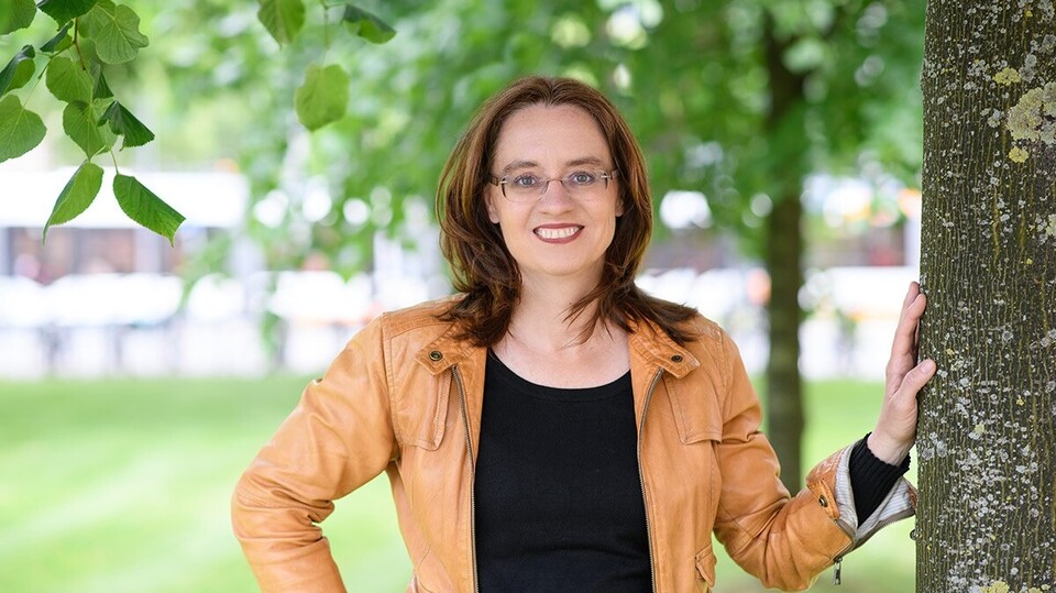 Prof. Michèle Tertilt, Ph.D Portrait von Michele Tertilt. Sie trägt einen schwarzen Pollover sowie eine orange farbige Jacke und steht auf einer Grünfäche.