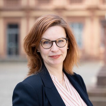 Sabine Gehrlein hat schulterlanges rotbraunes Haar. Sie trägt eine Brille, Perlenohrringe, eine Kette, ein rosé-weiß gestreiftes Hemd und ein dunkles Jackett. 
