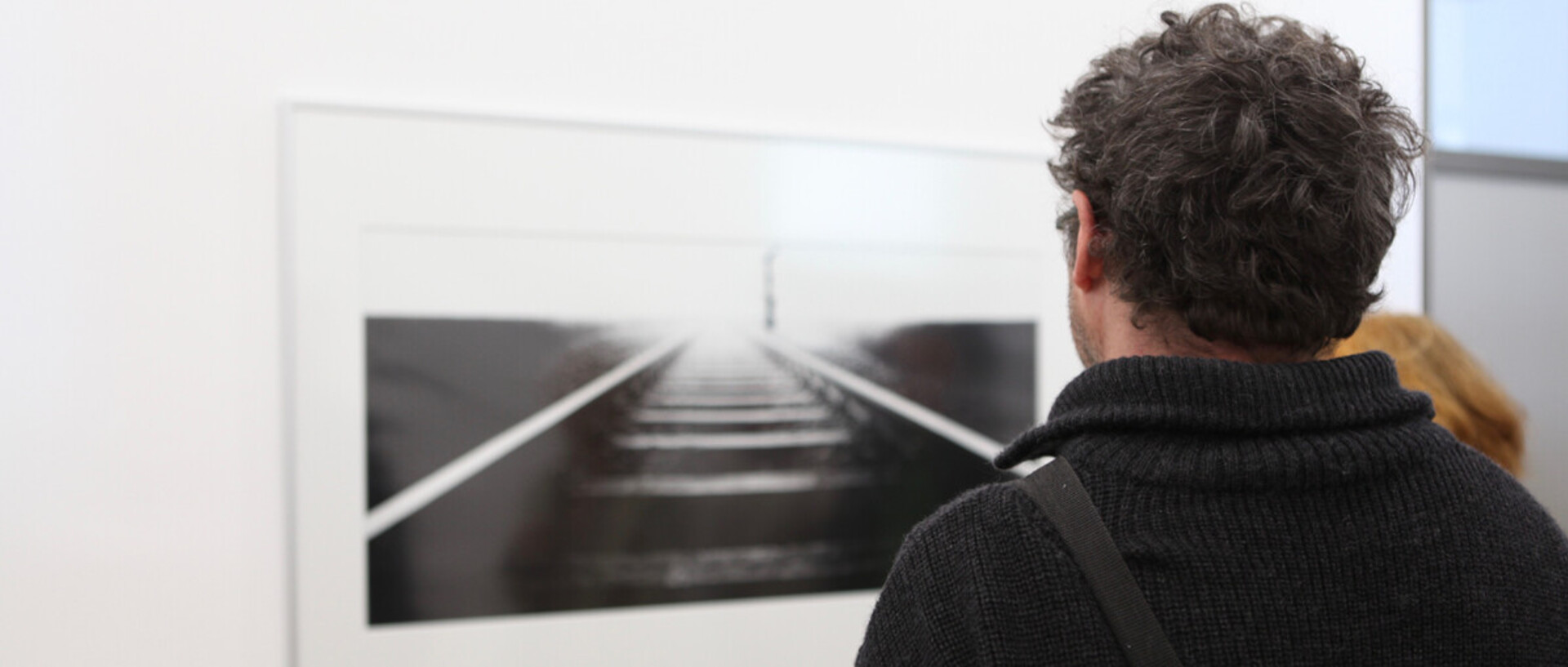 Ein Mann ist von hinten zu sehen. Er betrachtet ein Bild, das an einer weißen Wand hängt.