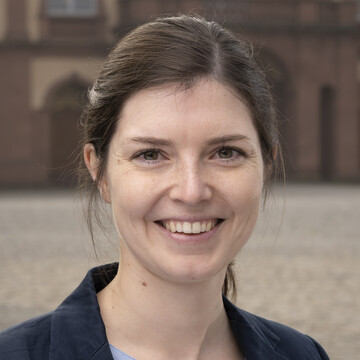 Janna Ried hat dunkle Haare und trägt eine hellblaue Bluse und einen dunklen Blazer. Im Hintergrund ist das Schloss zu sehen.