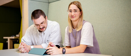 Ein Student und eine Studentin sitzen an einem Tisch. Der Student schaut auf einen Ipad vor sich, die Studentin direkt in die Kamera.