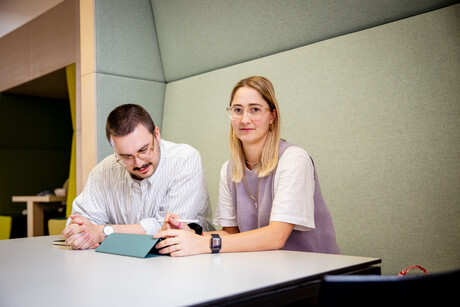 Ein Student und eine Studentin sitzen an einem Tisch. Der Student schaut auf einen Ipad vor sich, die Studentin direkt in die Kamera.