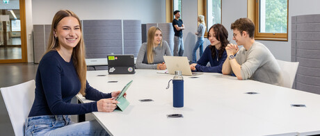 Eine Studentin sitzt an einem Arbeitstisch in B6. Im Hintergrund unterhalten sich 3 Studierende. 