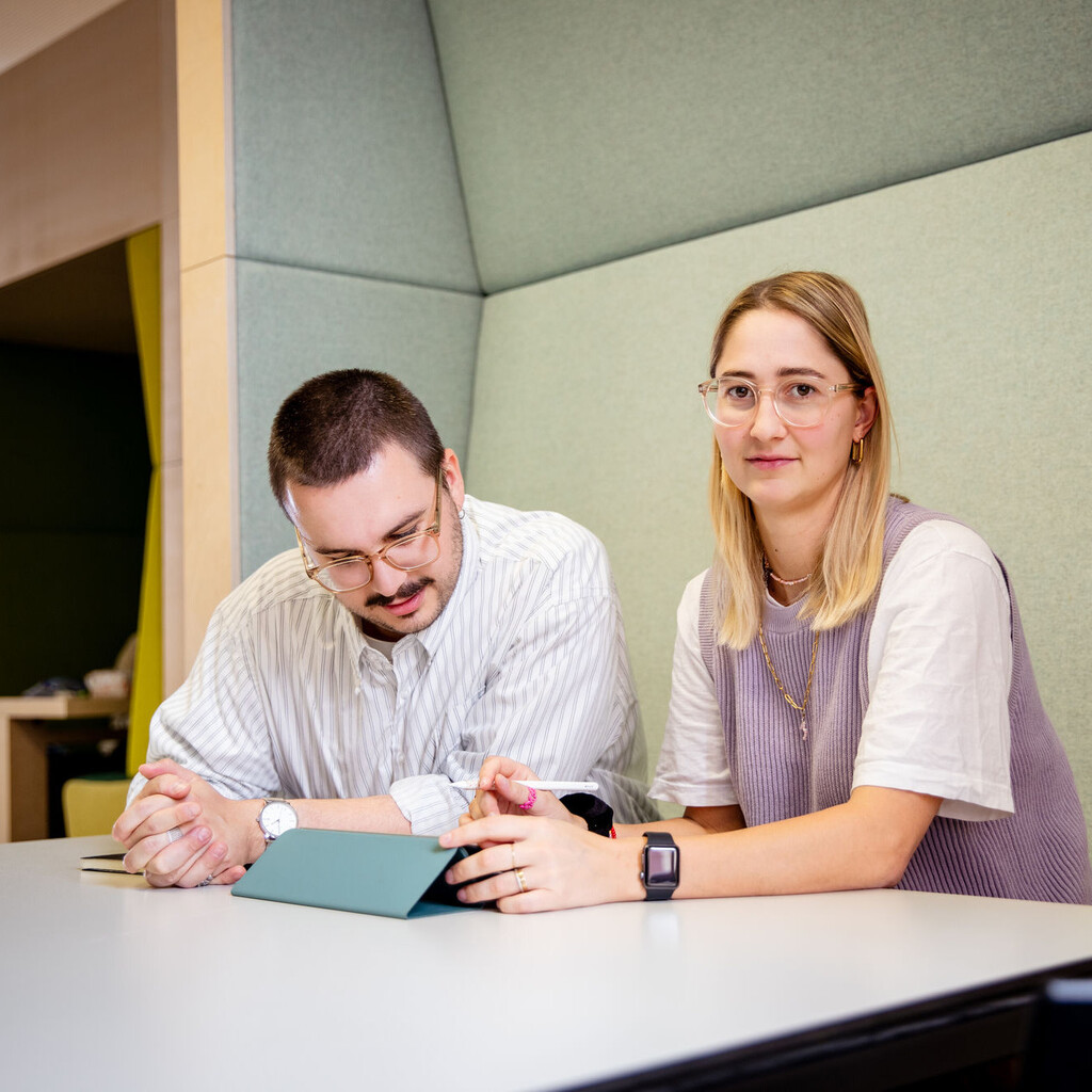 Ein Student und eine Studentin sitzen an einem Tisch. Der Student schaut auf einen Ipad vor sich, die Studentin direkt in die Kamera.