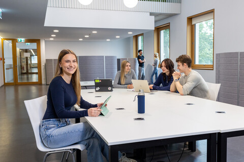 Eine Studentin sitzt an einem Arbeitstisch in B6. Im Hintergrund unterhalten sich 3 Studierende. 