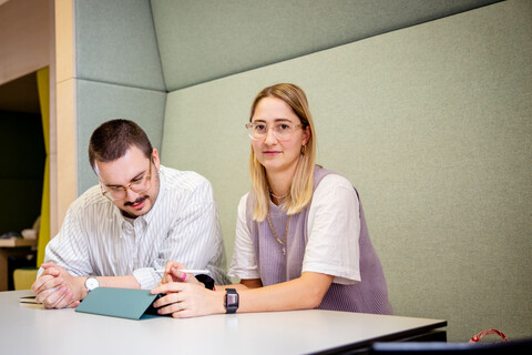 Ein Student und eine Studentin sitzen an einem Tisch. Der Student schaut auf einen Ipad vor sich, die Studentin direkt in die Kamera.
