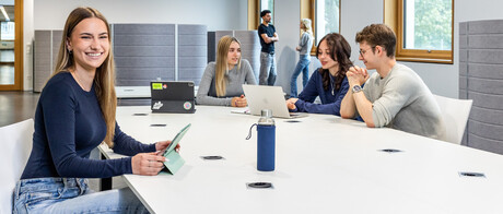 Eine Studentin sitzt an einem Arbeitstisch in B6. Im Hintergrund unterhalten sich 3 Studierende. 