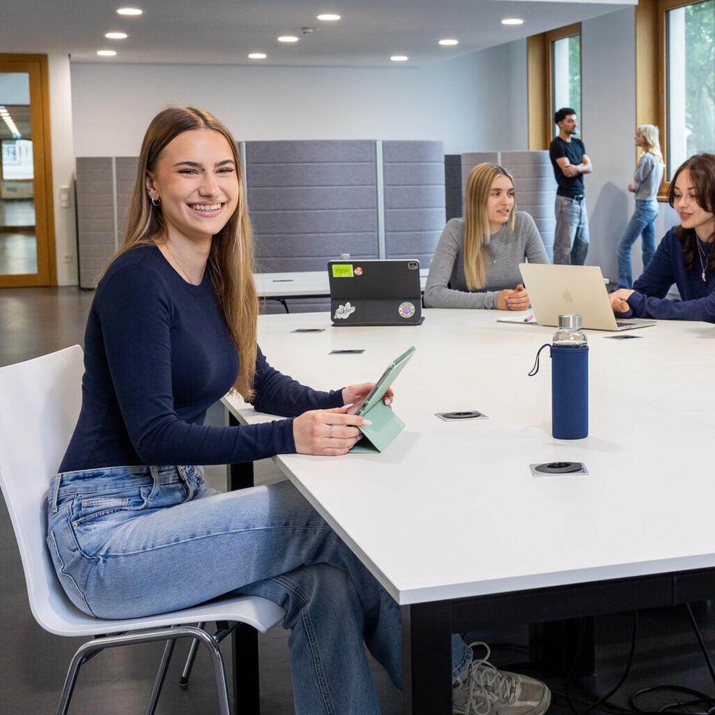 Eine Studentin sitzt an einem Arbeitstisch in B6. Im Hintergrund unterhalten sich 3 Studierende. 