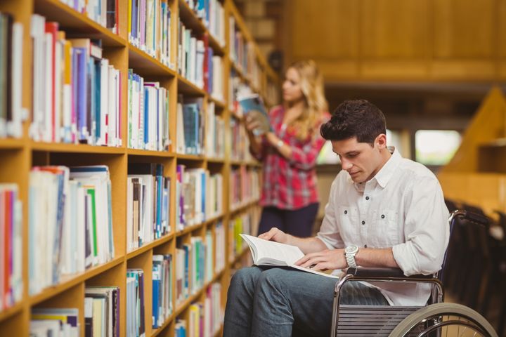 Mann im Rollstuhl liest vor einem Bücherregal in der Bibliothek etwas in einem Buch nach.