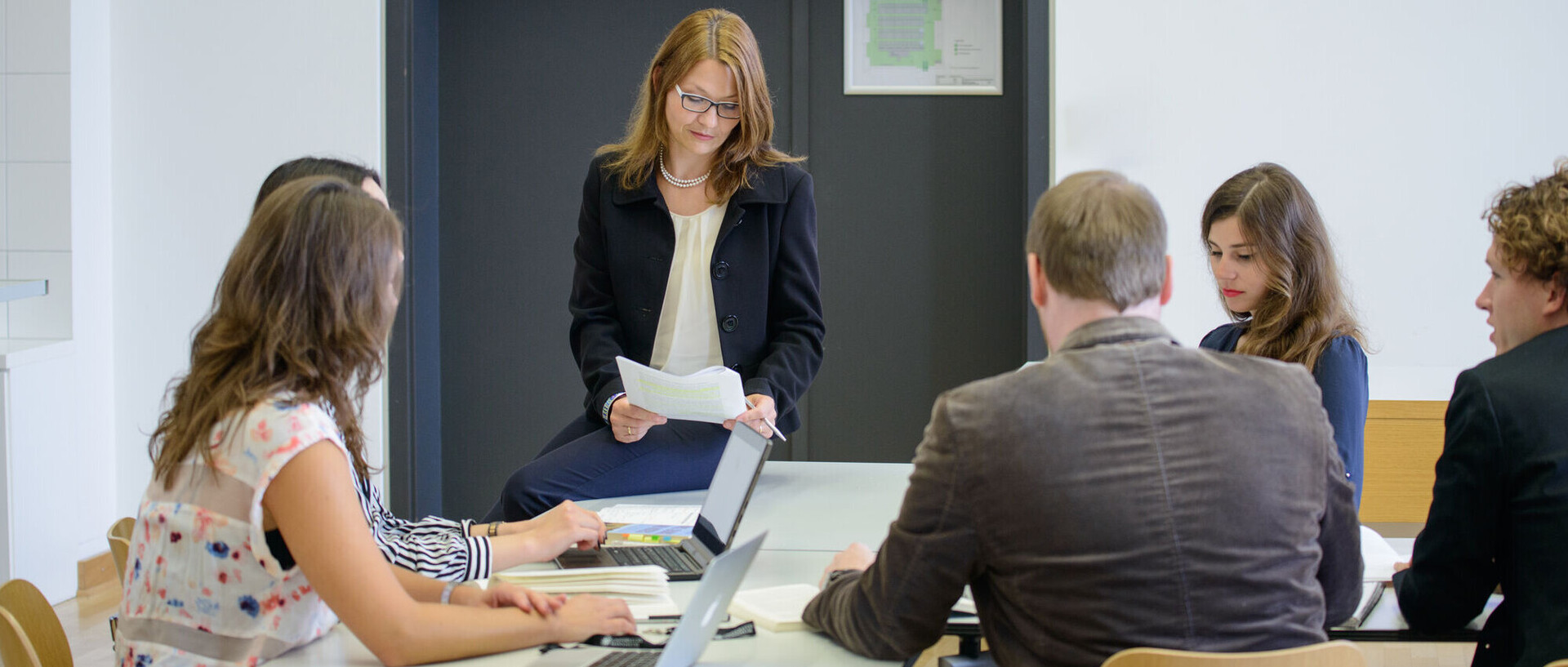 Five doctoral students sitting on tables in the seminar room while the teacher who also sits on a table reads a paper