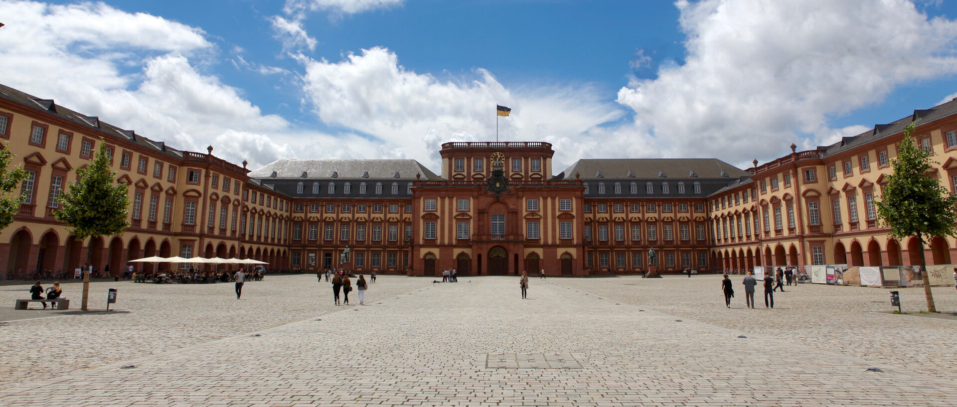 Das sandsteinfarbene Mannheimer Barockschloss mit Blick auf den Mittelbau bei strahlend blauem Himmel. Vereinzelt schlendern junge Menschen über das Kopfsteinpflaster auf dem Ehrenhof.