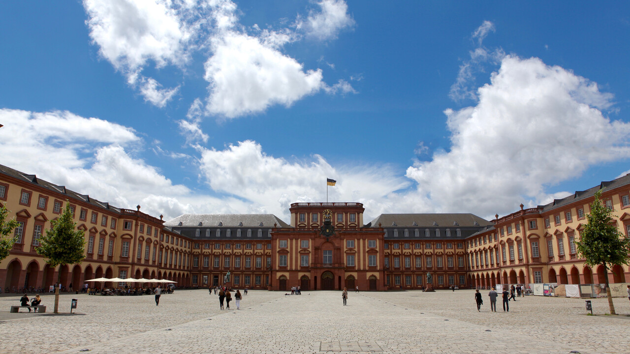 Das sandsteinfarbene Mannheimer Barockschloss mit Blick auf den Mittelbau bei strahlend blauem Himmel. Vereinzelt schlendern junge Menschen über das Kopfsteinpflaster auf dem Ehrenhof.