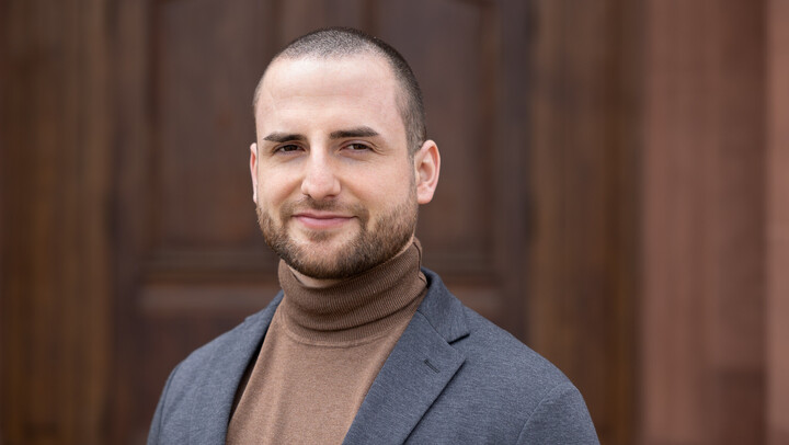 Yann Müller. He is wearing a grey jacket over a khaki sweater and is standing in front of a brown door.