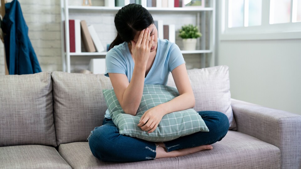 Eine Frau sitzt im schneidersitz auf dem Sofa. Auf ihrem Schoß liegt ein Kissen, sie fasst sich an die Stirn. 