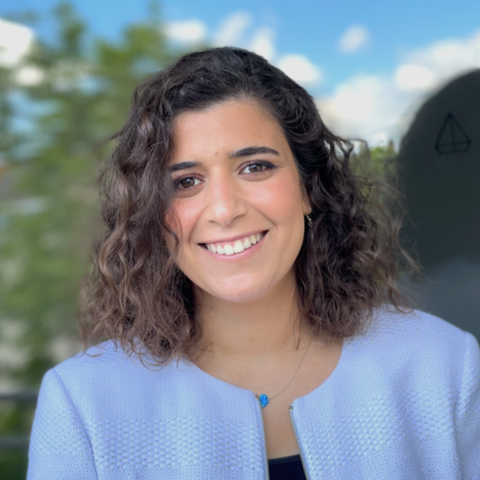 Amina trägt ein schwarzes Shirt unter ihrem fliederfarbenen Blazer. Ihre brauen Haare sind lockig und schulterlang. Sie lächelt und trägt Perlenohrringe.