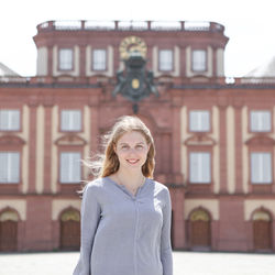 Anna Lehmann hat blonde lange Haare. Sie trägt eine graue Bluse und steht vor dem Mannheimer Schloss.