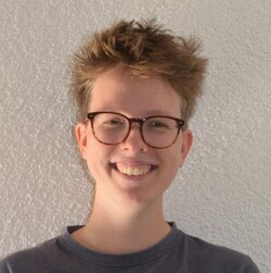 Anne Jaeschke has short blond hair and is smiling at the camera. She is wearing a blue T-shirt and brown horn-rimmed glasses.