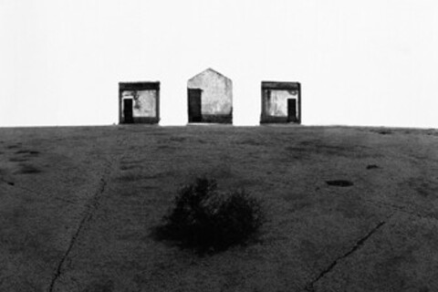 Schwarz-weiß Foto: Am Horizont sind zwei viereckige Hütten, dazwischen ist eine Hütte mit einem spitzen Dach.