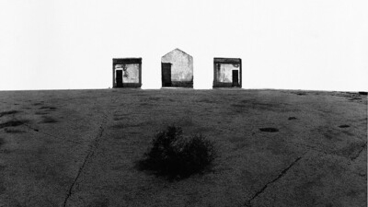 Schwarz-weiß Foto: Am Horizont sind zwei viereckige Hütten, dazwischen ist eine Hütte mit einem spitzen Dach.