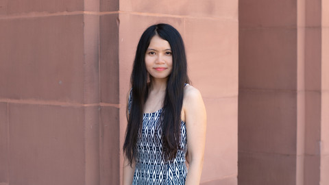 Hwee Bin Koh. She is wearing a black-white patterned dress and is standing in front of brown wall.