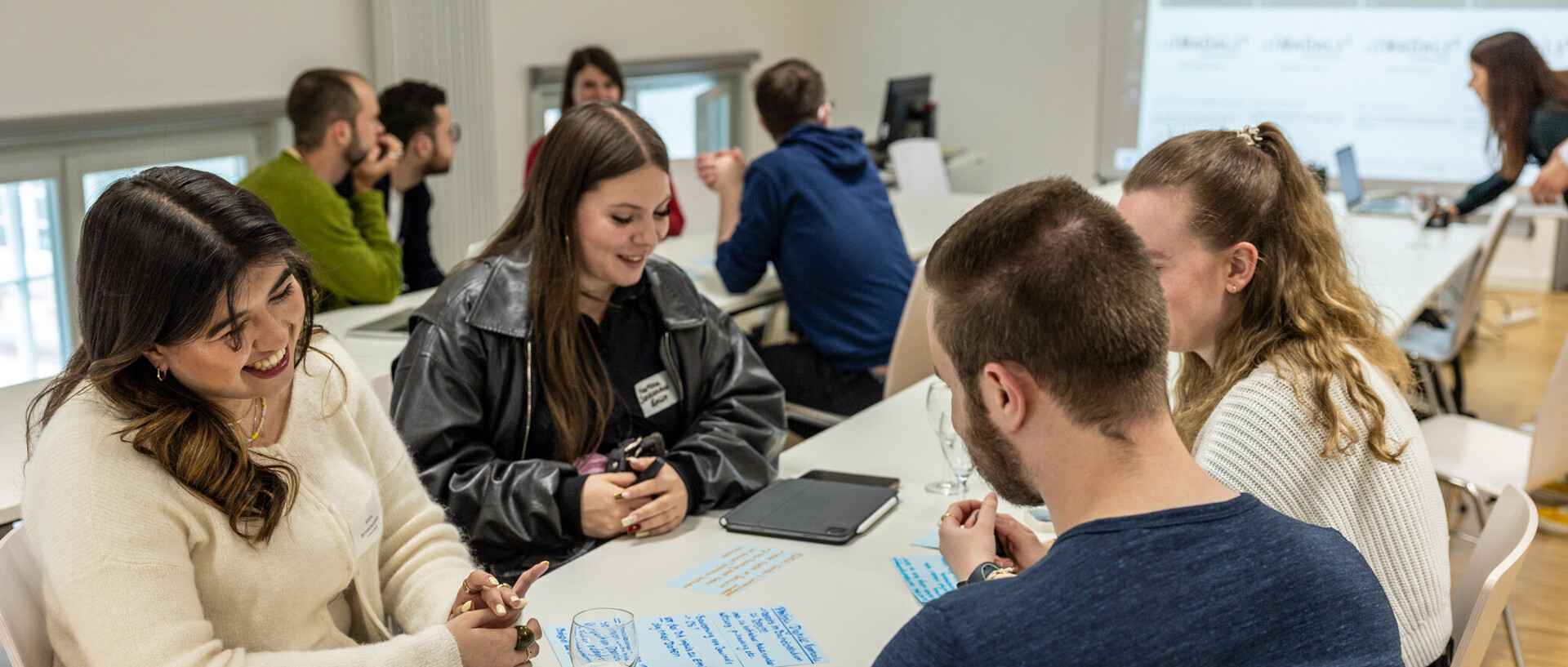 Workshop in hellem Seminarraum: Gruppen sitzen an Tischen und bearbeiten Aufgaben, eine Person im Vordergrund lächelt.