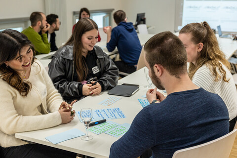 Workshop in hellem Seminarraum: Gruppen sitzen an Tischen und bearbeiten Aufgaben, eine Person im Vordergrund lächelt.