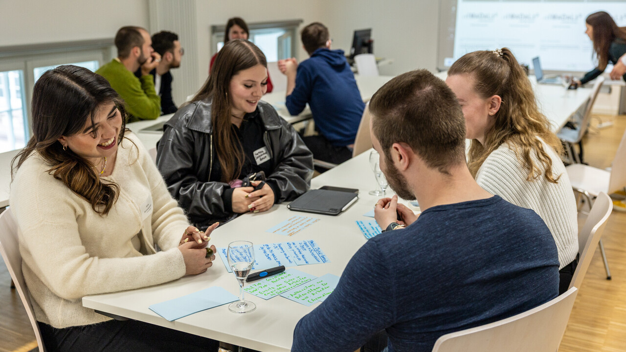 Workshop in hellem Seminarraum: Gruppen sitzen an Tischen und bearbeiten Aufgaben, eine Person im Vordergrund lächelt.