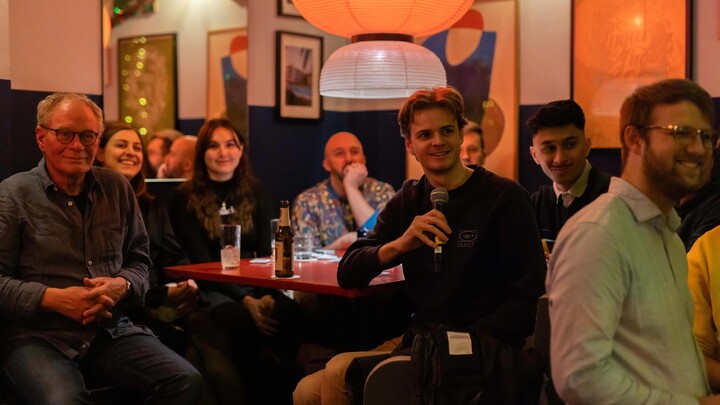 Mehrere Zuhörer*innen sitzen an einem Tisch in der Odeon Bar. Einer von ihnen spricht gerade in ein Mikrofon.