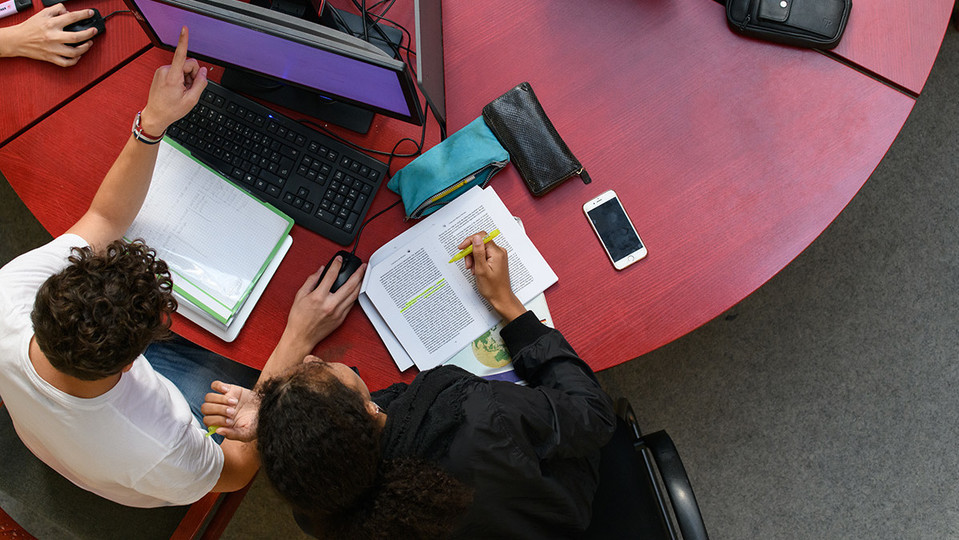 2 Studierende sitzen an einem runden roten Tisch mit Tastatur, Maus und Bildschirm sowie diversen darum verteilten Unterlagen und Schreibutensilien sowie einem Smartphone. Fotografiert wurde von oben. Der linke Studierende hält mit der rechten Hand die PC-Maus und deutet mit dem linken Zeigefinger auf den Bildschirm, auf den beide blicken. Der rechte Student hält einen Textmarker in der rechten Hand, unter der sich geheftete Papiere befinden, in deren oberster, aufgeschlagener Seite, bereits eine Passage gelb markiert ist. Seine linke Hand befindet sich neben seinem Kopf in einer Art, als ob er sich gerade dort kratzen wollte.