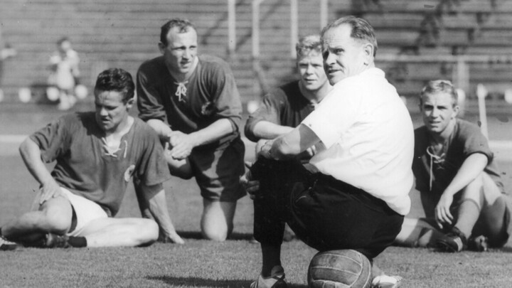 Schwarz-weiß Bild von Sepp Herberger auf dem Fußballplatz. Er trägt ein weißes Oberteil und sitzt auf einem Ball.