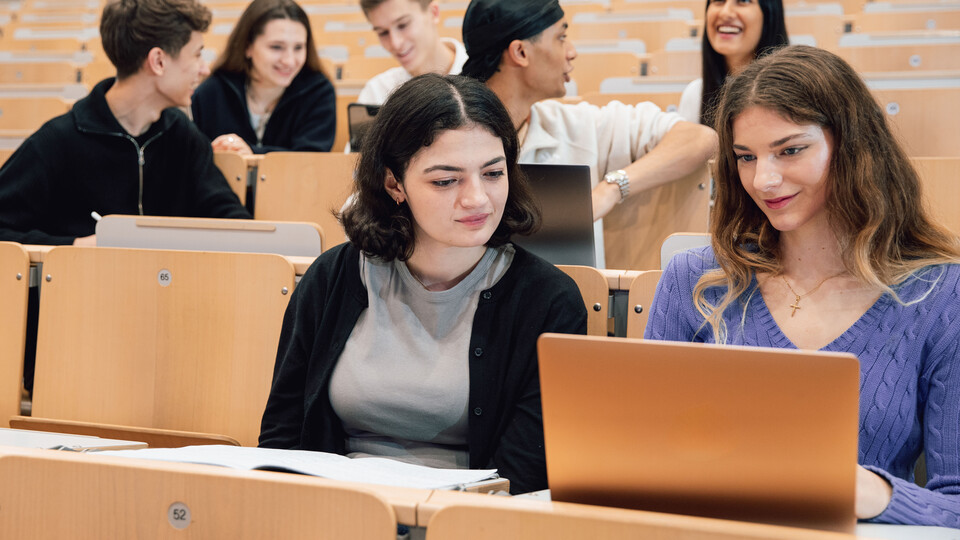 Studierende sitzen in einem Hörsaal. Sie unterhalten sich und schauen auf Laptops.