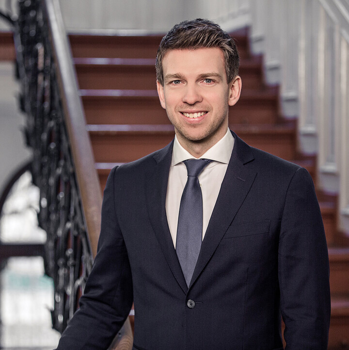 Portrait von Arnd Vomberg. Er trägt ein weißes Hemd, eine dunkelblaue Krawatte sowie einen dunkelblauen Blazer. Er steht in einem Treppenhaus.