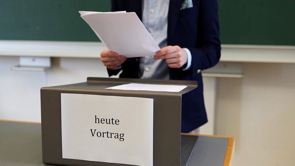 Im Hintergrund eine große grüne Tafel. Davor der mittlere Teil eines Körpers mit hellbelauem Hemd und dunkelblauem Jackett, der mit beiden Händen einen Stapel mit losen Papieren vor sich hält. Kopf und Beine sind nicht zu sehen. Wiederum davor ein Holztisch mit hellgrauer Oberfläche und Rednerpultaufsatz aus dunkelgrau gestrichenen Metall, auf dem weitere Papiere liegen. Auf der Vorderseite des Pultaufsatzes befindet sich ein großer weißer Zettel, auf dem in schwarzer Schrift flächenfüllend "heute Vortrag" gedruckt ist.