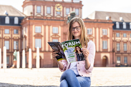 Studentin sitzt auf einer Bank auf dem Ehrenhof und liest in der aktuellen Ausgabe des FORUM-Magazins