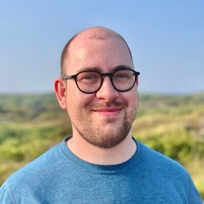 Christopher Klamm vor einer Wiese und blauem Himmel im Sonnenschein. Er trägt eine Brille und einen blauen Pullover.